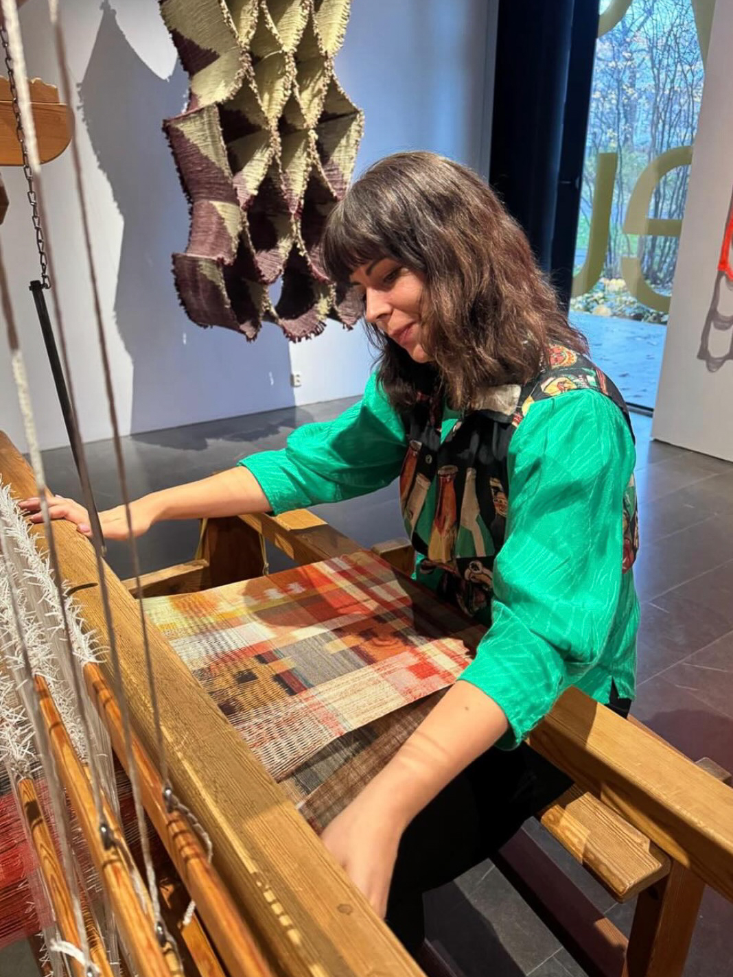 Maneka Ghosh sitter vid en trävävstol i en ljus gallerimiljö. Hon bär en grön skjorta med mönstrad krage och lutar sig framåt med handen på vävstolen. I bakgrunden hänger ett skulpturalt textilverk i lila och beige. Dagsljus syns genom ett fönster till höger.