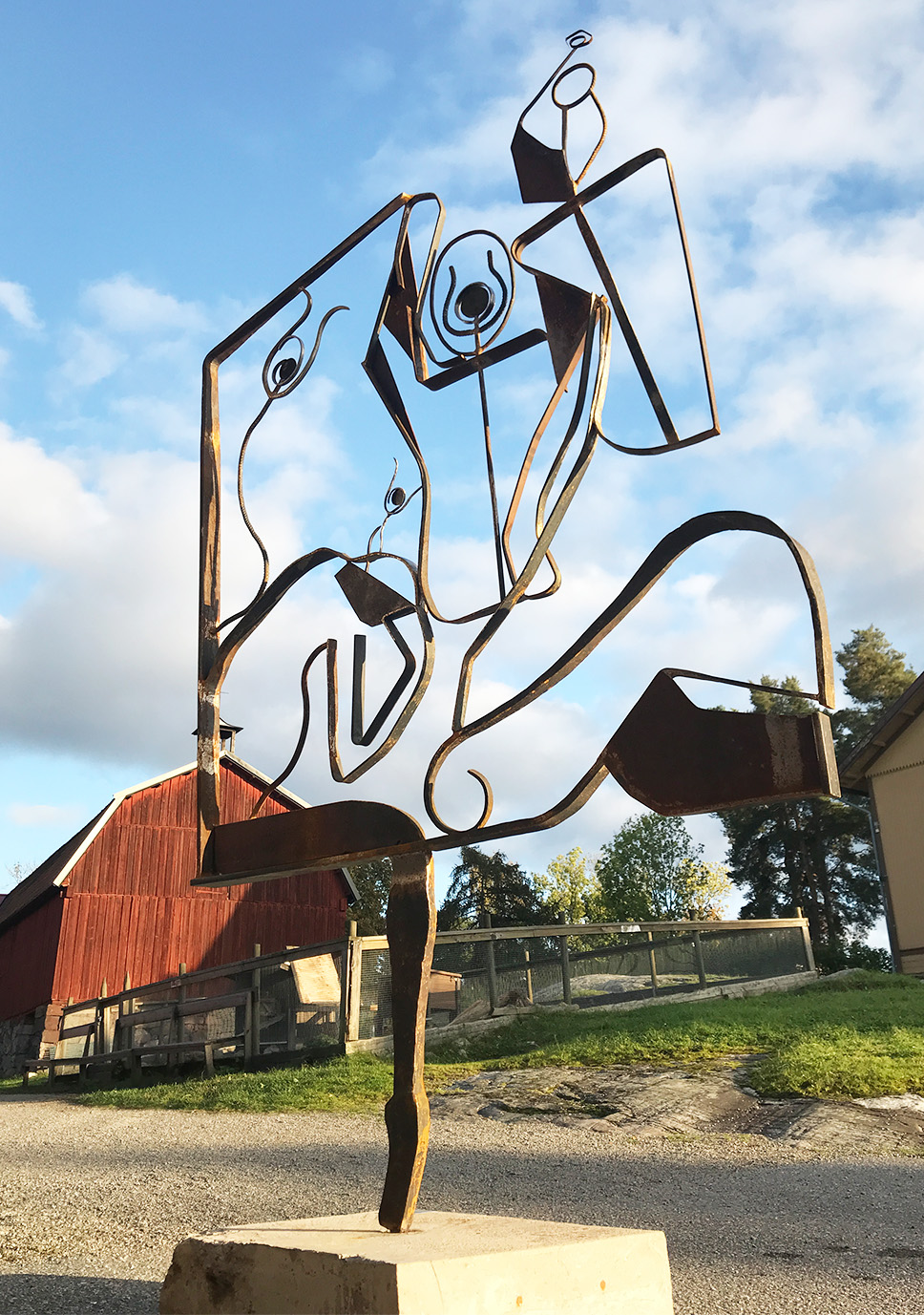 En stor fristående järnskulptur på en betongbas, fotograferad utomhus i solljus. Skulpturen är gjord av smidda och svetsade järnelement som bildar abstrakta, dynamiska former inspirerade av kropp och rörelse. Det rostbruna järnet kontrasterar mot en blå himmel. I bakgrunden syns en röd ladugård och gröna träd.