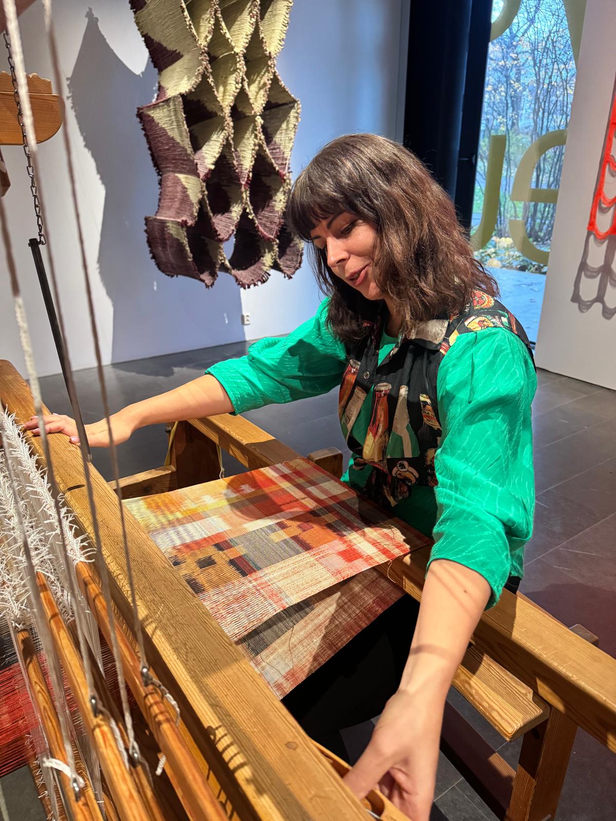 Maneka Ghosh arbetar vid en trävävstol i en gallerimiljö med högt i tak. Hon bär en grön skjorta med mönstrad krage och lutar sig engagerat framåt med båda händerna på vävstolen. Väven har ett rutmönster i röda, orange och grå toner. I bakgrunden hänger ett skulpturalt beige och lila textilverk och ett fönster med utsikt mot blommande träd syns.