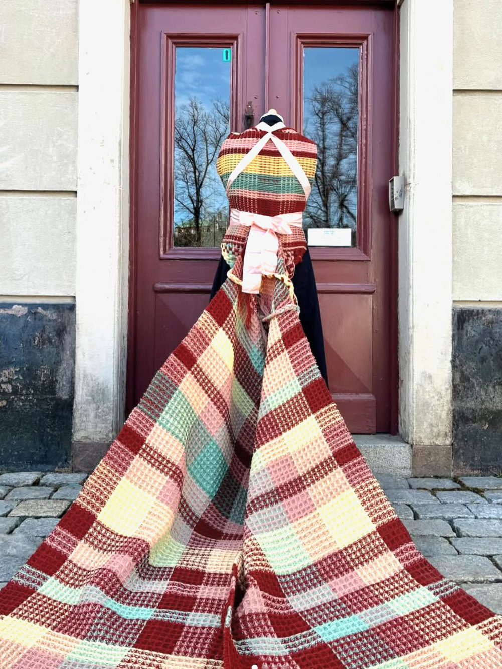 Ett handvävt textilverk i våffelstruktur draperat på en svart skyltdocka utan huvud, placerad framför en vinröd pardörr. Verket har ett rutmönster i rosa, rött, grönt, gult och beige och breder ut sig ned mot kullerstensmarken.